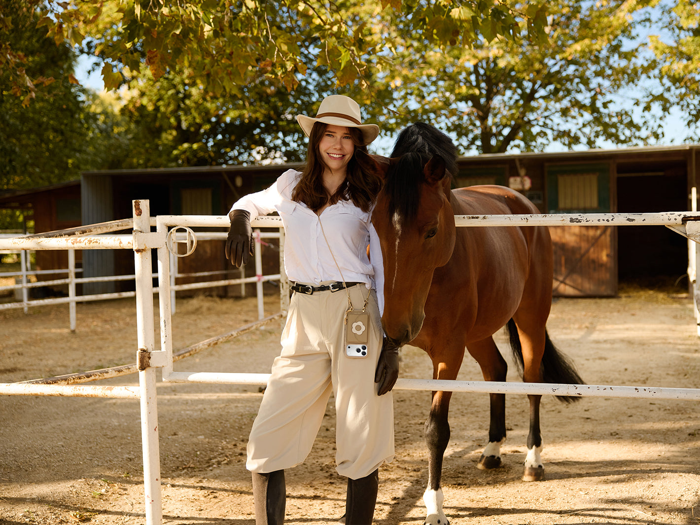 Woman wearing a taupe chain crossbody phone case beside a horse at an autumn stable