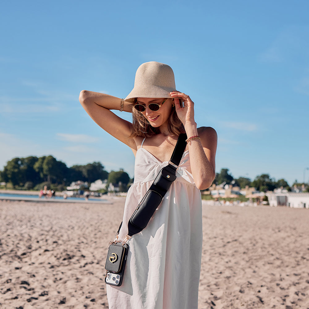 A woman on a sandy beach wearing a beige straw hat, sunglasses, a white sleeveless dress, and a black crossbody bag with a phone pouch.
